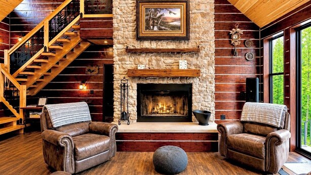 L’intérieur d’une cabane en rondins, avec une cheminée en pierre, des canapés en cuir, un escalier en bois et une grande fenêtre.