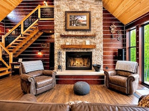 A log cabin interior with a stone fireplace, leather sofas, a wooden staircase, and a large window.