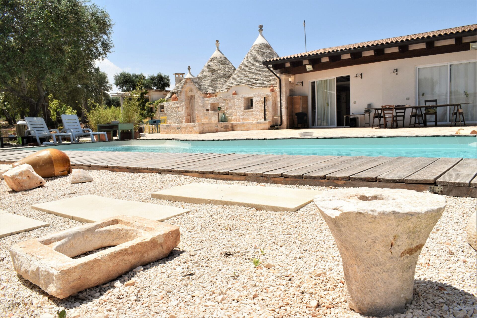 Un área de piscina con tumbonas de piedra, una terraza de madera y un edificio con techos cónicos al fondo.