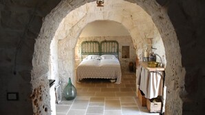 Une chambre à coucher rustique avec une arche en pierre, un lit doté d’une tête de lit en métal vert et une petite table sur laquelle se trouvent une lampe et un bol.