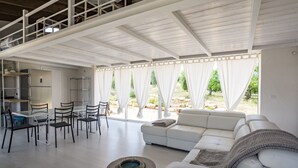 Un vaste espace de vie comprenant un canapé blanc, une table à manger avec des chaises et un coin cuisine équipé d’un réfrigérateur et d’étagères.