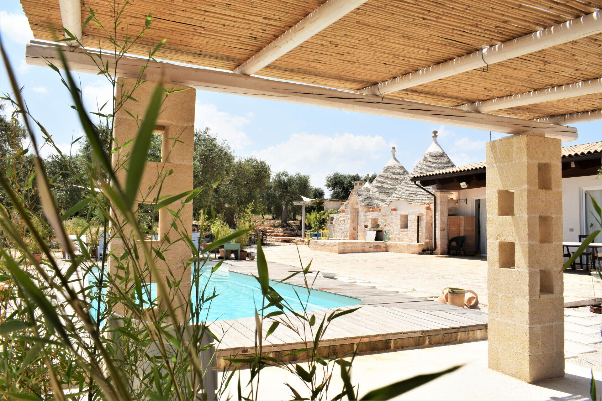 Un espace piscine agrémenté d’une pergola, des bâtiments en pierre en arrière-plan et des plantes de bambou au premier plan.
