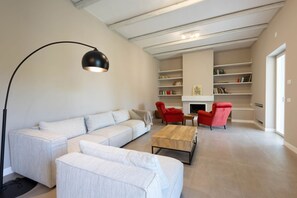 Ein modernes Wohnzimmer mit einer Eckcouch, einem Couchtisch und roten Sesseln. Im Hintergrund sind Bücherregale und ein Kamin zu sehen.