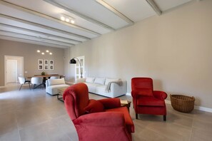 A spacious living area with a white sofa, red armchairs, a dining table, and a kitchen in the background.