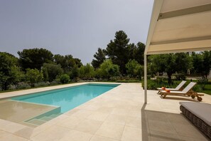 Piscina retangular com deck revestido, rodeada por vegetação exuberante e árvores. Duas cadeiras de descanso com almofadas e um toldo.