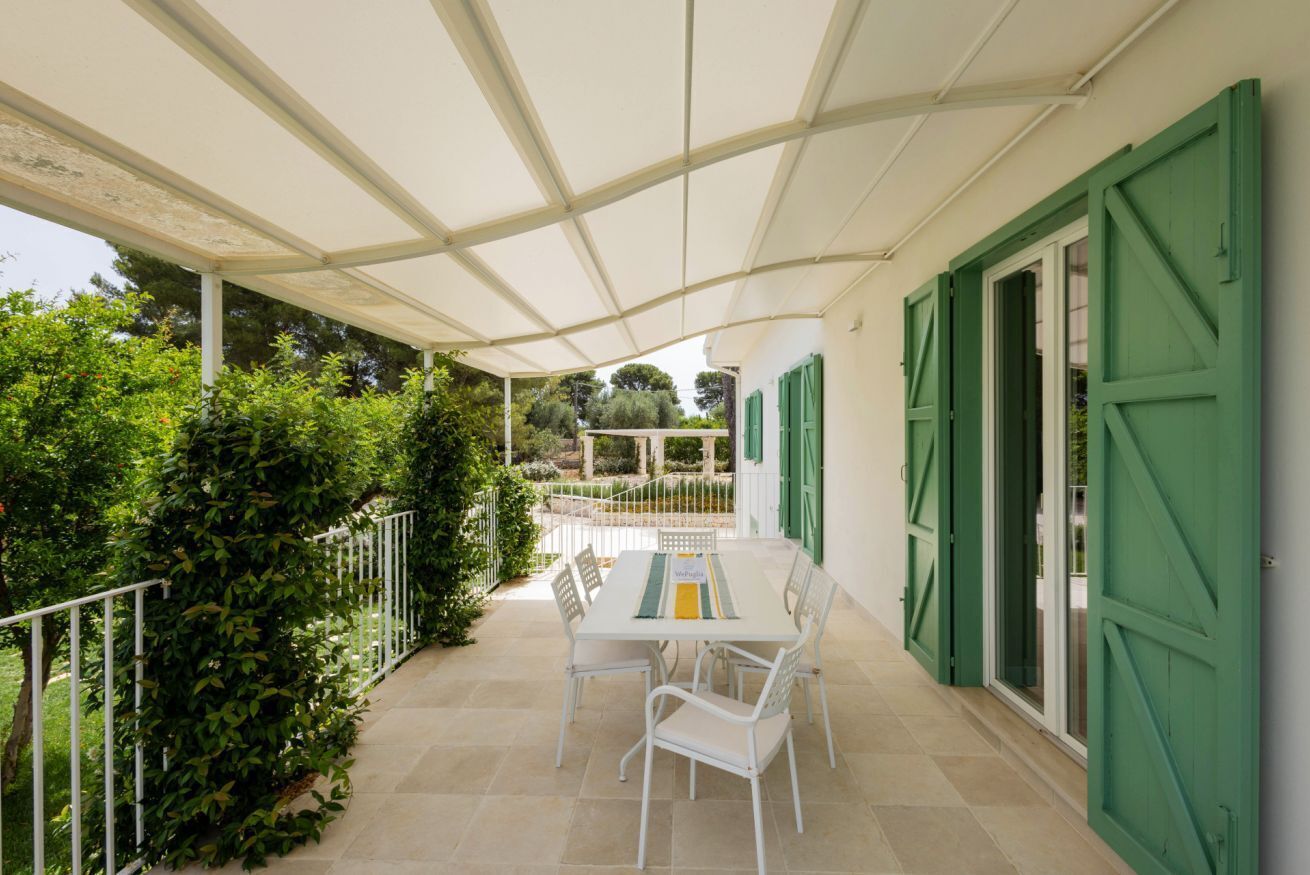 Eine überdachte Terrasse mit Tisch und Stühlen, umgeben von Pflanzen und einem weißen Zaun.