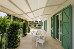 A covered patio with a table and chairs, surrounded by greenery and a white fence.