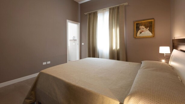 Een hotelkamer met een bed, een nachtkastje met een lamp, een schilderij aan de muur en een raam met gordijnen.