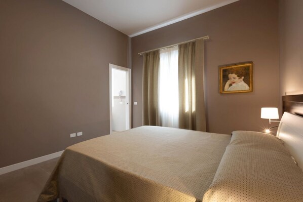 A hotel room with a bed, a nightstand with a lamp, a painting on the wall, and a window with curtains.