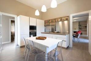Uma cozinha moderna com armários brancos, uma ilha central e uma área de jantar com cadeiras transparentes.