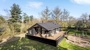 Une cabane moderne dotée d’une grande terrasse, entourée d’arbres et d’une zone herbeuse.
