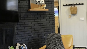 Una habitación con pared de ladrillo negro, un televisor montado, un sillón de cuero amarillo con una manta gris, una cesta tejida y una obra de arte enmarcada.