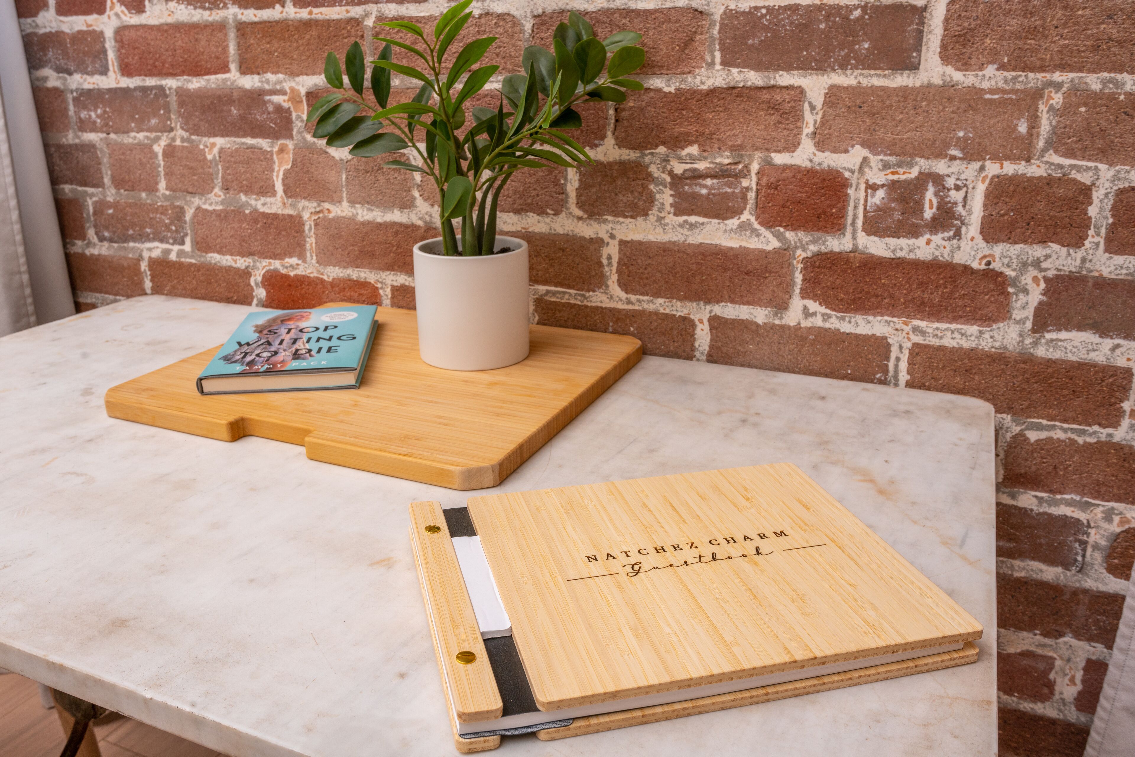 Een houten snijplank met een boek getiteld 'Act of Waiting' en een potplantje op een marmeren tafel, voor een bakstenen muur.