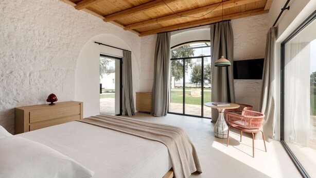 A bedroom with a large bed, wooden ceiling, a small table with chairs, and a view through large windows.