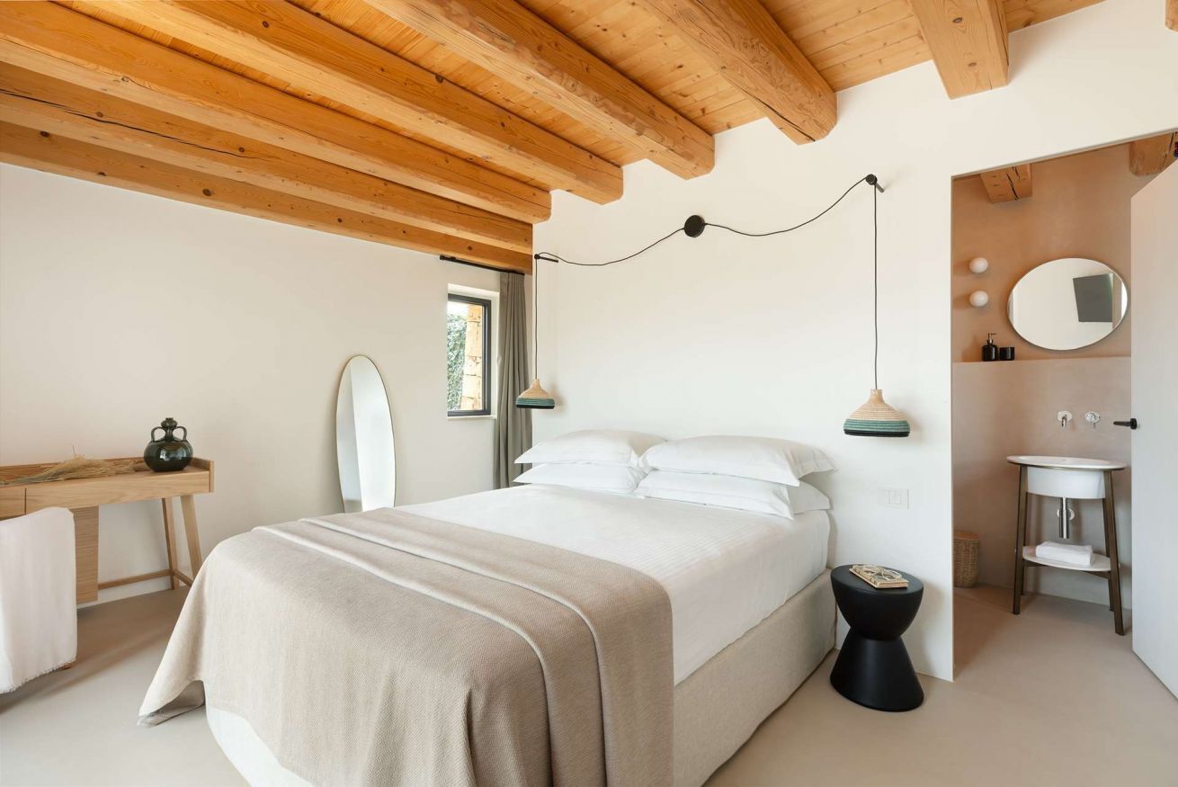 Een slaapkamer met een bed, nachtkastje en een klein krukje. Er is een raam met gordijnen, een spiegel en een wandlamp.