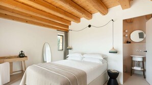 A bedroom with a bed, bedside table, and a small stool. There is a window with curtains, a mirror, and a wall-mounted light fixture.