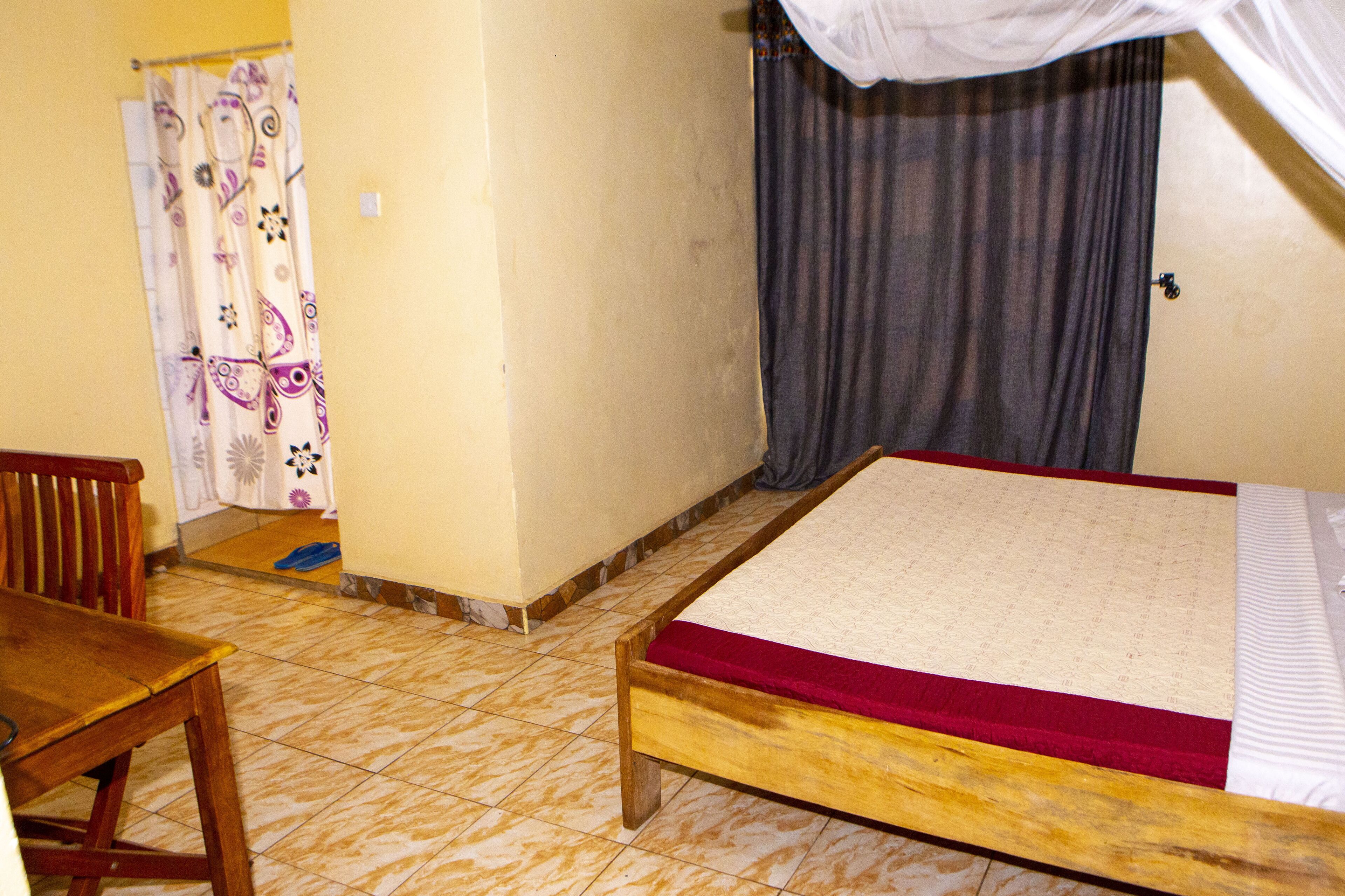 A room with a wooden bed, a wooden chair, a small table, a curtain with a paisley pattern, and a tiled floor.