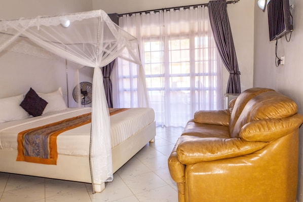 A bedroom with a bed, a leather sofa, a bedside fan, and a window with curtains. - Nv Fort Lugard Hotel Iganga (Iganga)