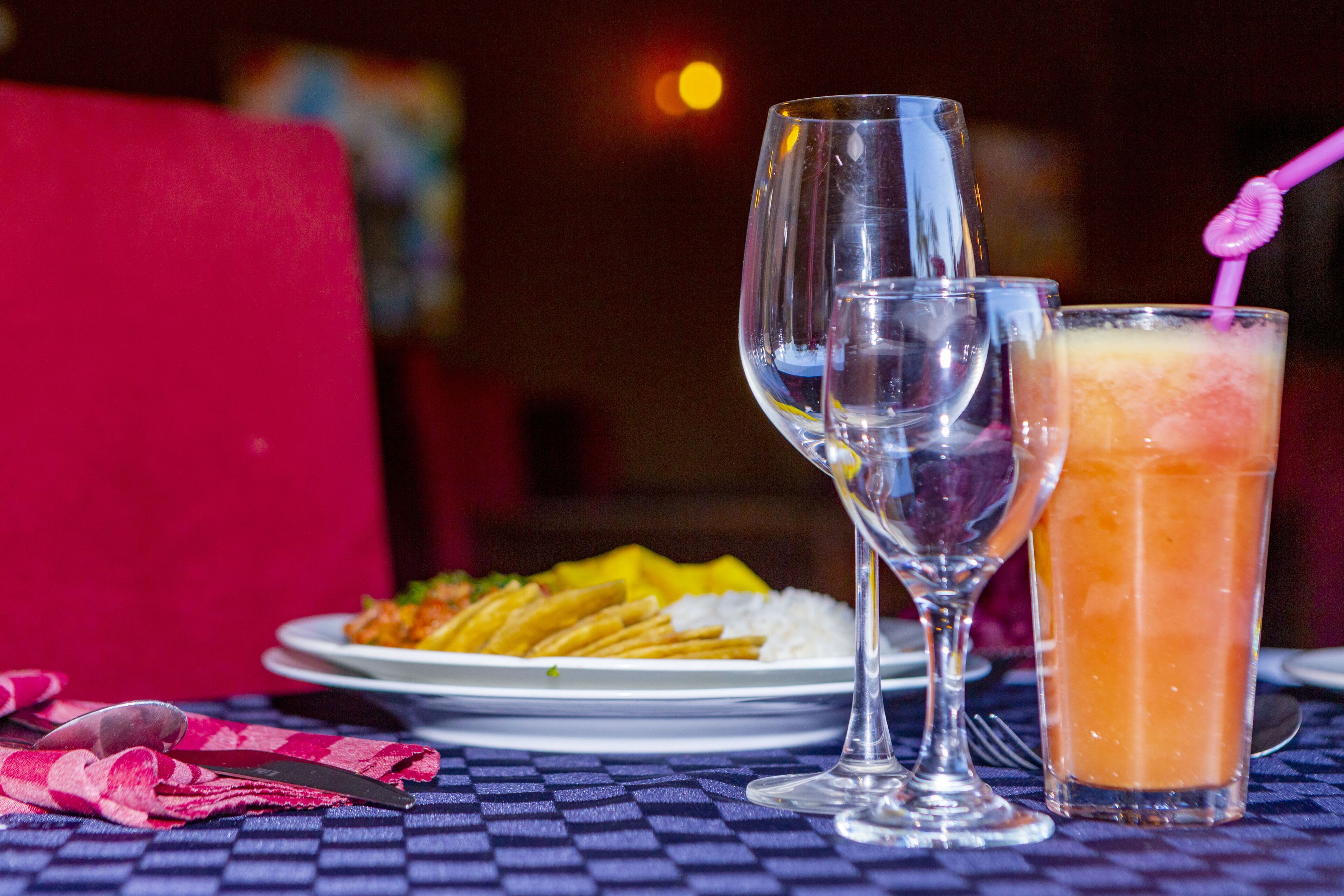 A dining table with a plate of food, two wine glasses, and a tall glass of orange beverage.