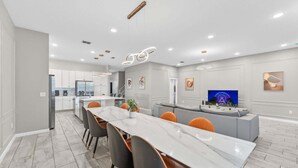 A modern kitchen with white cabinets, a central island, and a dining area with a long table and chairs. - Huge Family Home! Sleeps 26! (Kissimmee)