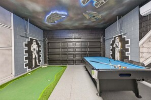 Uma mesa de bilhar com tema Star Wars, uma área com grama sintética verde e uma porta de garagem.