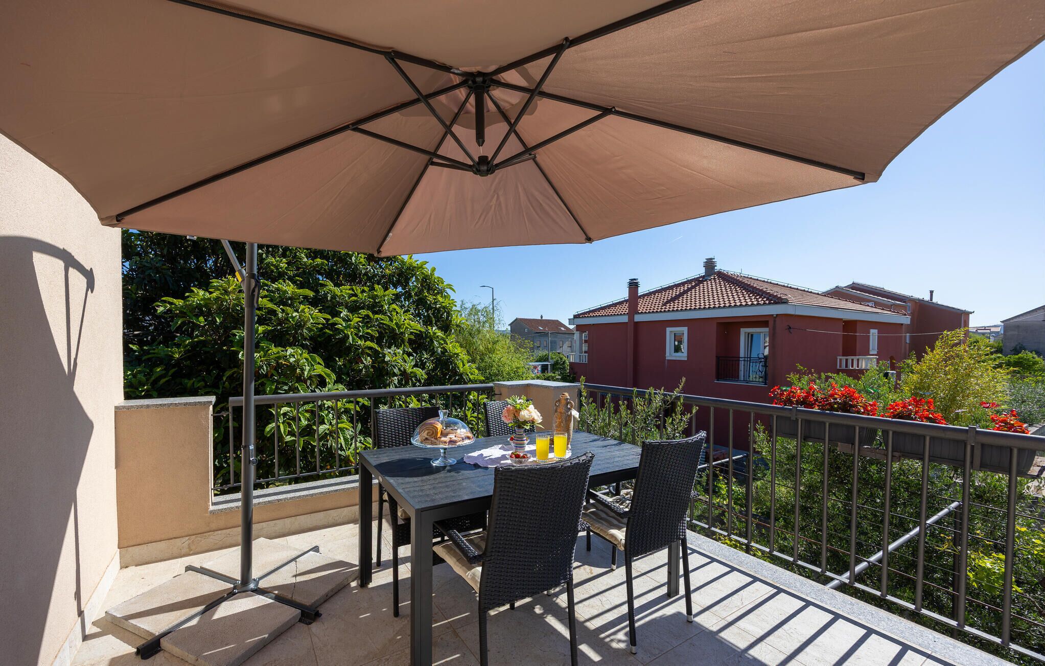 Uma varanda com mesa e cadeiras, um grande guarda-sol e vista para um edifício vermelho ao fundo.