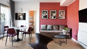 Una sala moderna con una pared de acento roja, una mesa redonda de comedor con sillas, un sofá y una pequeña mesita de noche con una planta.