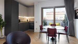 Una cocina moderna con armarios oscuros, una zona de comedor con una mesa redonda y sillas rojas, y un gran ventanal con vistas a edificios y una planta en el exterior.