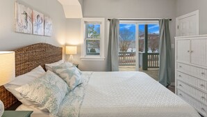 Een slaapkamer met een groot bed, een rieten hoofdbord, een nachtkastje met een lamp, een raam met gordijnen en uitzicht op het omliggende landschap.