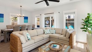 A modern living room with a sectional sofa, a dining area with wicker chairs, and a large window with curtains. - Luxury OBX 3BR, Private Hot Tub, Near the Beach (Corolla)