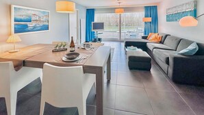Una sala de estar moderna con comedor, sofá y televisor. Hay un cuadro en la pared y una ventana con cortinas.