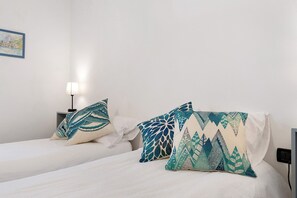 A hotel room with two beds, each with a white bedspread and decorative pillows. There is a nightstand with a lamp and a wall-mounted power outlet.