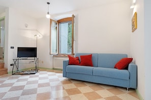 Un salon avec un canapé bleu, des coussins rouges, un téléviseur posé sur un meuble, une fenêtre et un radiateur.