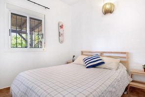 Une chambre à coucher avec un lit, une fenêtre, un luminaire fixé au mur et une petite table sur laquelle se trouve un vase.