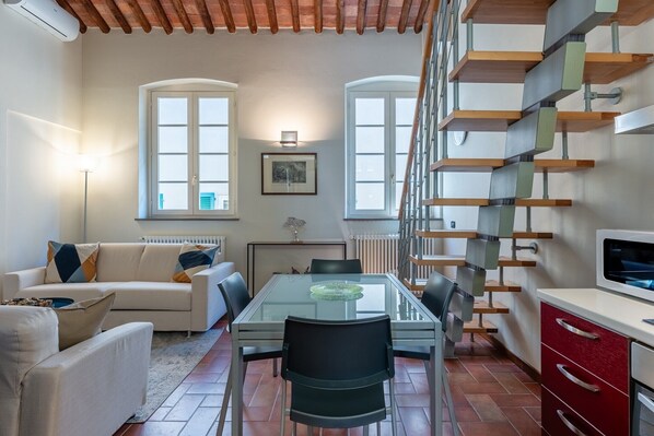 A modern living room with a glass dining table, a sofa, a staircase, and a kitchen area.
