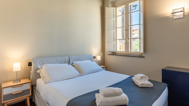 A neatly made bed with white linens, a bedside lamp, and a window with curtains. - Toro's Home Lucca Apartment with Wi-Fi and Air Conditioning (Lucca)