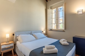 A neatly made bed with white linens, a bedside lamp, and a window with curtains.