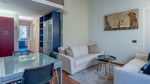 A living room with a glass dining table, a white sofa, a blue armchair, and a painting on the wall. - Toro's Home Lucca Apartment with Wi-Fi and Air Conditioning (Lucca)