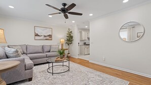 Ein Wohnzimmer mit grauem Sofa, einem runden Couchtisch, Deckenventilator, Stehlampe, einem Blumentopf und einer Küche im Hintergrund.