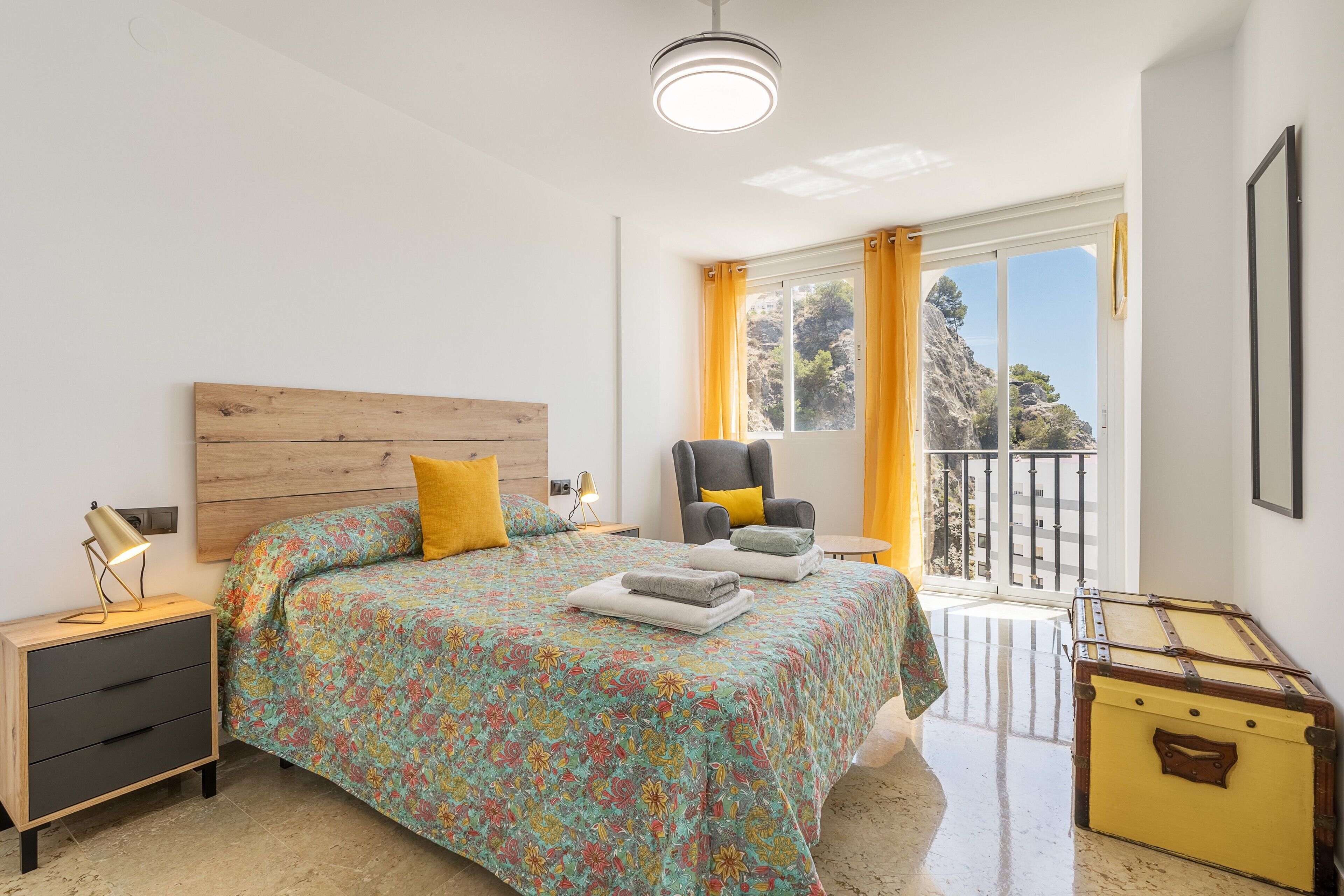 A bedroom with a wooden headboard, a bed with floral bedding, a nightstand with a lamp, a chair with a cushion, and a suitcase.