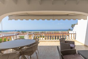 A covered patio with a table and chairs, a view of the sea, and a balcony with a railing.