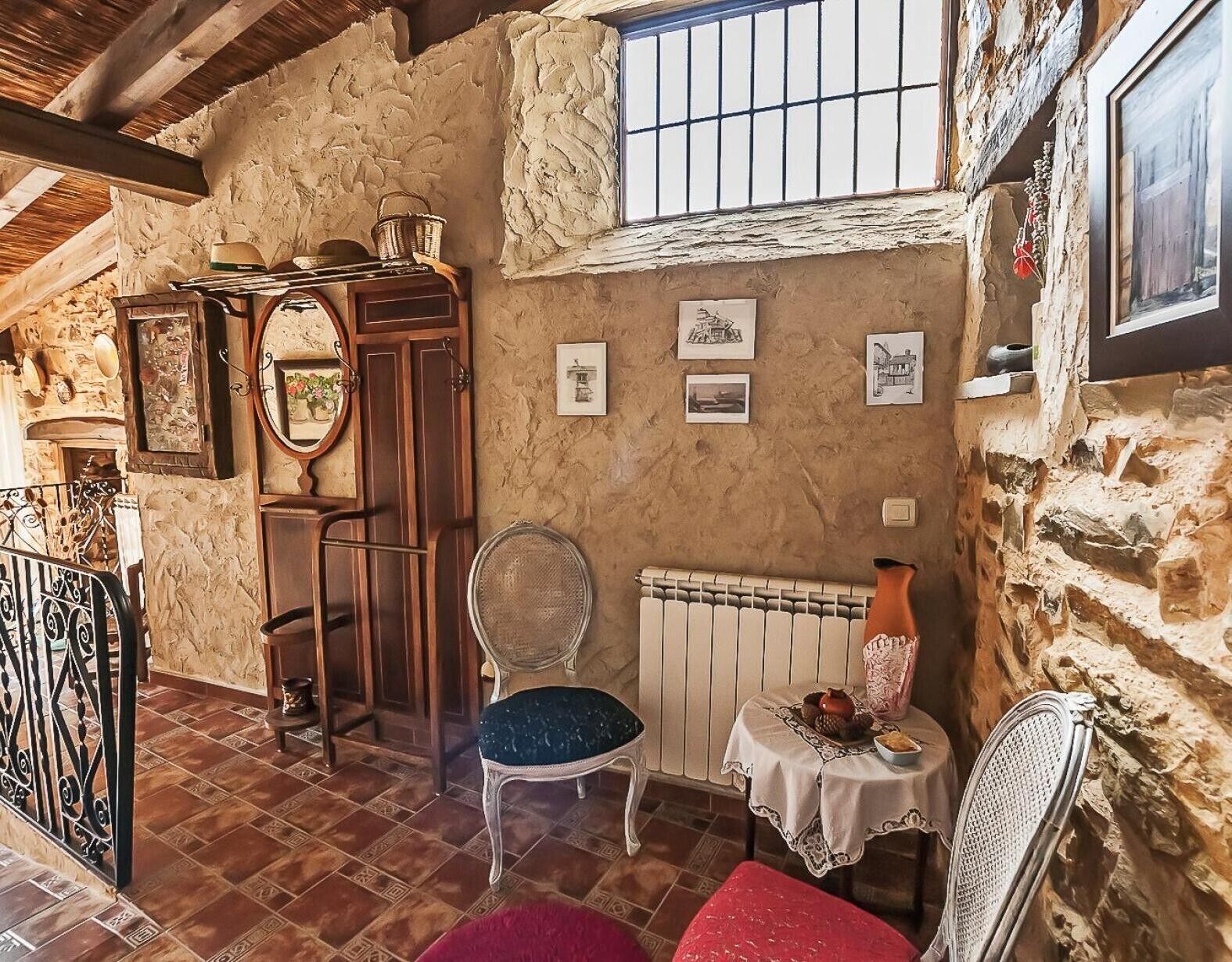 Een rustieke kamer met stenen muren, een houten deur, een radiator, een stoel, een tafel met een vaas en een raam met tralies.