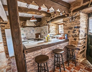 A rustic kitchen with stone walls, a tiled backsplash, wooden beams, and a bar area with stools.