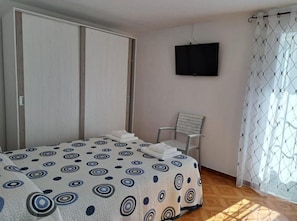 Une chambre à coucher avec un lit, une chaise, une armoire, un téléviseur et une fenêtre avec des rideaux.