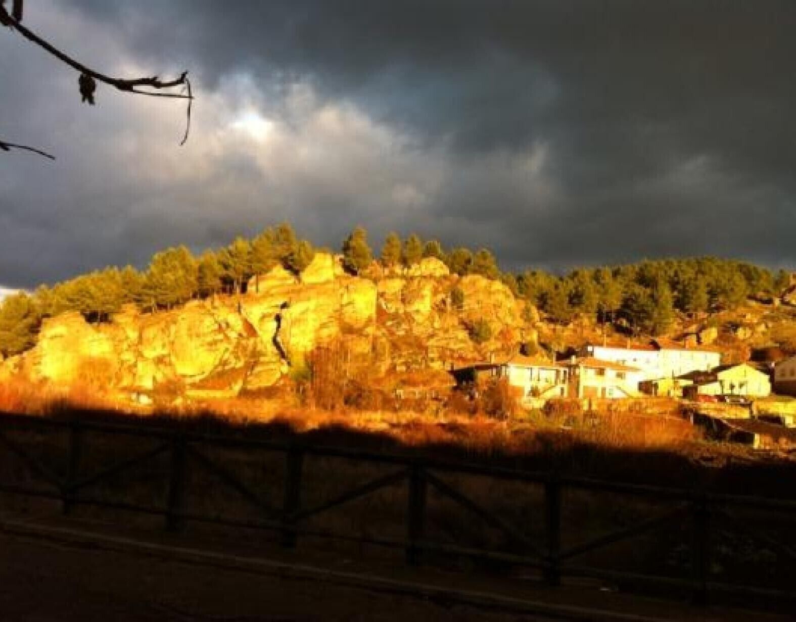 Paisaje rural con una colina rocosa, una valla y algunos edificios.