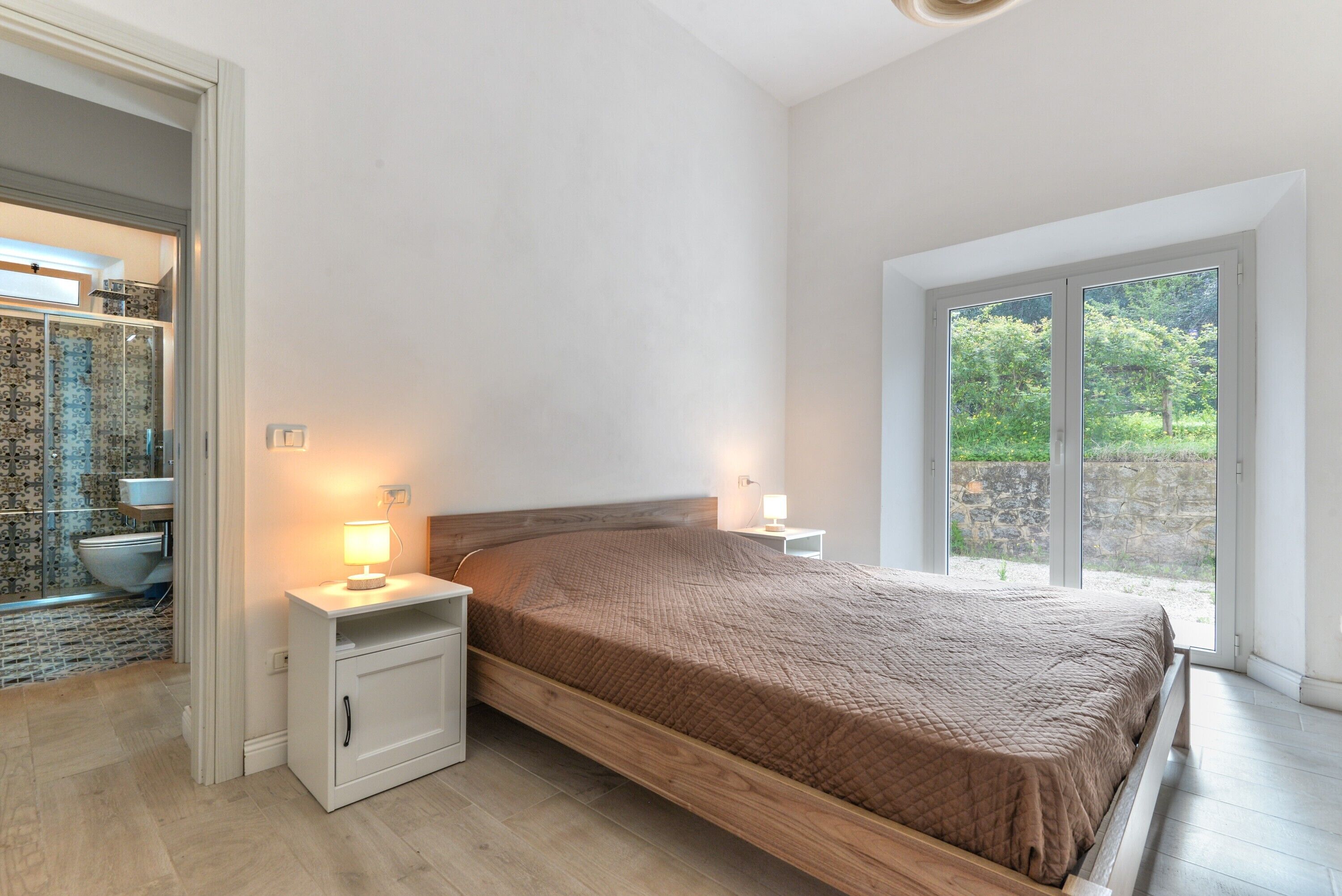 A bedroom with a bed, a nightstand, a bedside table, a bathroom with a toilet, and a view of greenery through a large window.
