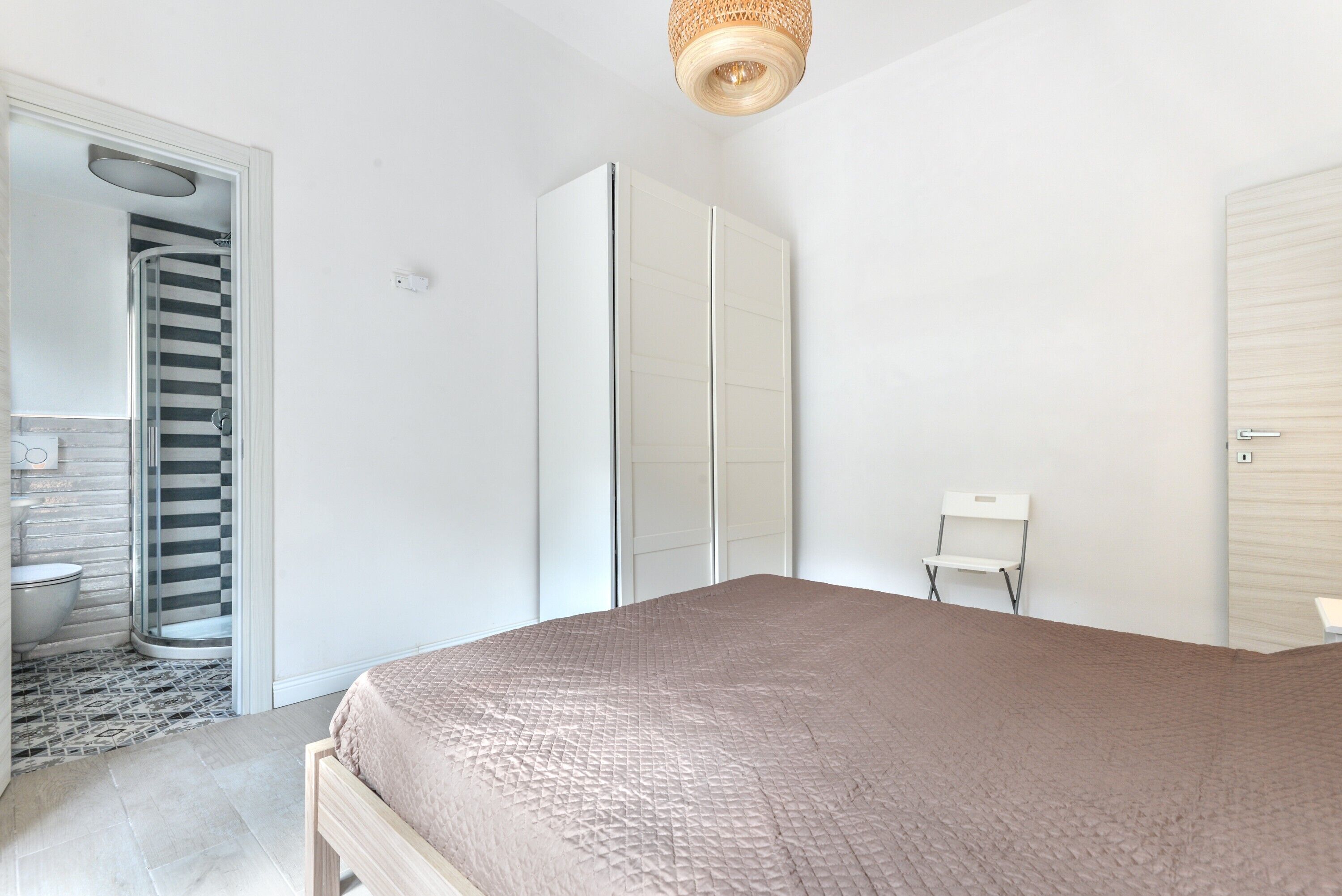 A bedroom with a bed, a wardrobe, a chair, and a bathroom with a shower.