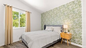 Ein Schlafzimmer mit einem Bett, einem Nachttisch, einem Fenster mit Vorhängen und einer Wand mit einem Blattmuster.