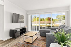 A modern living room with a flat-screen TV, a sofa, a coffee table, and a large window with blinds. - The Concord Place A3 (Peoria)
