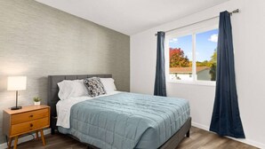 Un dormitorio con cama, mesita de noche, lámpara y cortinas.